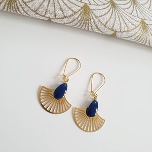 以下が含まれることがあります： ダークブルーのしずく型アクセントが付いた、金色の扇形のイヤリングのペア。 イヤリングはフック留め式で、背景に模様のある生地が入った白い表面に表示されています。 イヤリングはジュエリーアイテムです。