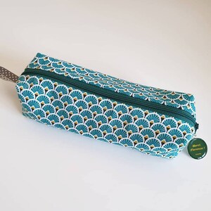 Può includere: Un astuccio per matite verde e oro con chiusura a zip e un motivo a forma di ventaglio. L'astuccio ha un piccolo cartellino verde attaccato con il testo "Merci Florence!"