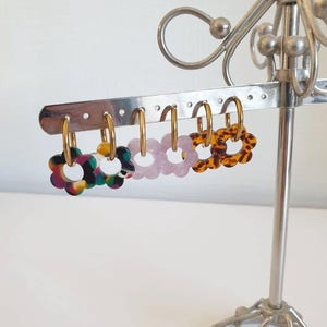 Può includere: Esposizione di orecchini a cerchio a forma di fiore in vari colori, tra cui multicolore, giallo, lilla e tartarugato. Gli orecchini sono esposti su un espositore argentato.