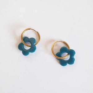 Puede incluir: Un par de pendientes de aro dorados con colgantes de flores de color verde azulado. Los colgantes de flores tienen un diseño de margarita. Los pendientes se presentan sobre un fondo blanco.