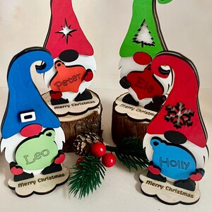 Puede incluir: Cuatro adornos de gnomos navideños de madera. Cada gnomo tiene un sombrero de diferente color: rojo, verde, azul y rojo. Cada gnomo sostiene un letrero en forma de corazón con un nombre: Peter, Ela, Leo y Holly. La base de cada gnomo dice "Merry Christmas".