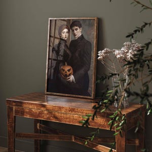 May include: A framed print featuring a vintage-style portrait of a couple holding a carved jack-o'-lantern. The artwork is in a wooden frame and placed on a wooden table. The color palette is dark, with a focus on black, brown, and orange.