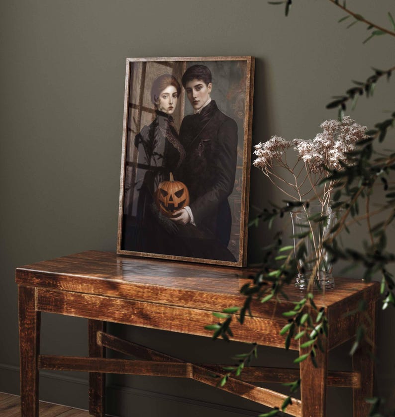 May include: A framed print featuring a vintage-style portrait of a couple holding a carved jack-o'-lantern. The artwork is in a wooden frame and placed on a wooden table. The color palette is dark, with a focus on black, brown, and orange.
