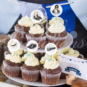 May include: A two-tiered white stand holding chocolate cupcakes with white frosting and blue sprinkles. Decorative circular toppers feature images of a fedora, black shoes, white boots, a microphone, and a person in a military-style jacket.