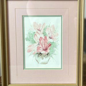 May include: Watercolor painting of pink and red lilies in a vase, framed in gold. The artwork is set against a pink mat and a light green border. The painting is a floral still life.