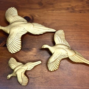 May include: Three gold-colored bird sculptures in flight, with detailed wing and feather designs. The birds are arranged on a dark wooden surface, creating a decorative display. The sculptures vary in size, adding visual interest.