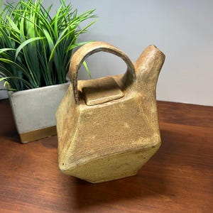 May include: A handcrafted, geometric teapot with a tan and brown finish. The teapot features a rectangular body, a curved handle, and a small, flat lid. The teapot is made of ceramic or stoneware and is a unique piece of kitchenware.