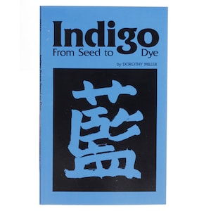 Könnte beinhalten: Ein blaues Buch mit dem Titel "Indigo From Seed to Dye" von Dorothy Miller. Der Umschlag zeigt ein schwarzes Quadrat mit einem stilisierten japanischen Schriftzeichen in Weiß.