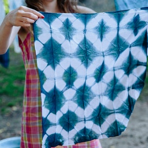 An indigo workshop student holds out and displays a lattice patterned bandana. Fuzzy white stripes crisscrossing a blue field. The color is an aqua teal that will oxidize to pure indigo after being dyed in Graham Keegans Indigo and Shibori dye kit