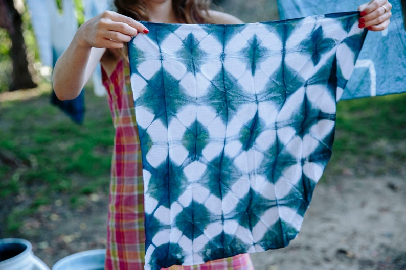 An indigo workshop student holds out and displays a lattice patterned bandana. Fuzzy white stripes crisscrossing a blue field. The color is an aqua teal that will oxidize to pure indigo after being dyed in Graham Keegans Indigo and Shibori dye kit