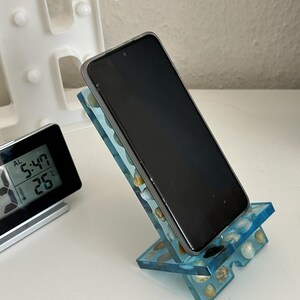 Könnte beinhalten: Ein Smartphone auf einem blauen Handyhalter aus Kunstharz, in den kleine, helle Muscheln eingearbeitet sind. Der Halter besteht aus zwei ineinandergreifenden Teilen, die das Gerät in einem Winkel zum Betrachten halten, mit einer klaren Oberfläche.