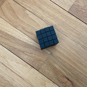 May include: A black cube made of smaller cubes, each with a smooth, flat surface. The cube is sitting on a wooden surface.