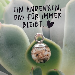 Könnte beinhalten: Ein kugelförmiger Anhänger mit einer silberfarbenen Metallkappe und einer Öse, gefüllt mit kleinen, bunten Sand- und Muschelfragmenten. Der Text "EIN ANDENKEN, DAS FÜR IMMER BLEIBT." mit einem schwarzen Herzsymbol steht über dem Anhänger.
