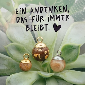 Könnte beinhalten: Drei kugelförmige Anhänger mit Metallösen, die Haare und Blattgoldflocken enthalten. Der Text lautet: EIN ANDENKEN, DAS FÜR IMMER BLEIBT. Die Anhänger haben unterschiedliche Größen, zwei mit goldfarbenen und einer mit silberfarbenen Metallteilen.