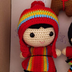 Puede incluir: Una muñeca de crochet con un gorro y un vestido a rayas rojas y arcoíris. La muñeca tiene el pelo negro, la piel blanca y ojos grandes negros.