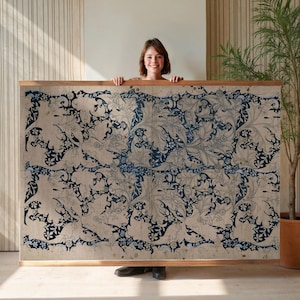 Puede incluir: Un gran textil rectangular con un patrón repetitivo de motivos florales y de hojas azules sobre un fondo beige. El textil está enmarcado en un marco de madera marrón claro. Una persona sostiene el textil.