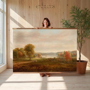 May include: A large framed landscape painting featuring a serene outdoor scene with trees, a lake, and a cloudy sky. The artwork is held by a person in front of a wooden wall. The frame is a warm brown color.