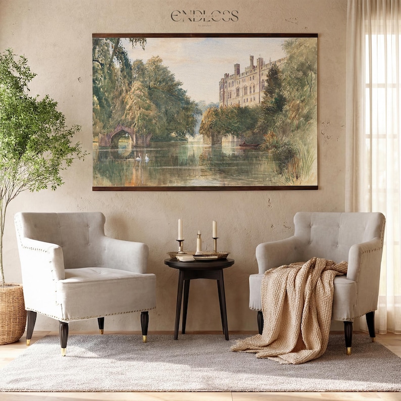 May include: A watercolour landscape print showing a lake scene with a bridge and a large building. Two light grey armchairs with dark legs and gold tips are positioned near a small black side table with candles. A beige throw is draped over one chair.