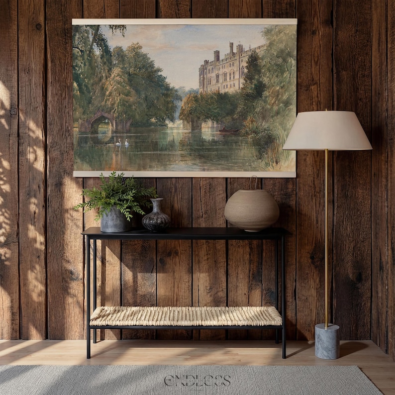May include: A watercolour painting of a castle and lake is displayed above a black console table with a woven shelf. A floor lamp with a cream shade and a marble base stands to the right. Decorative vases and a potted plant are on the table.