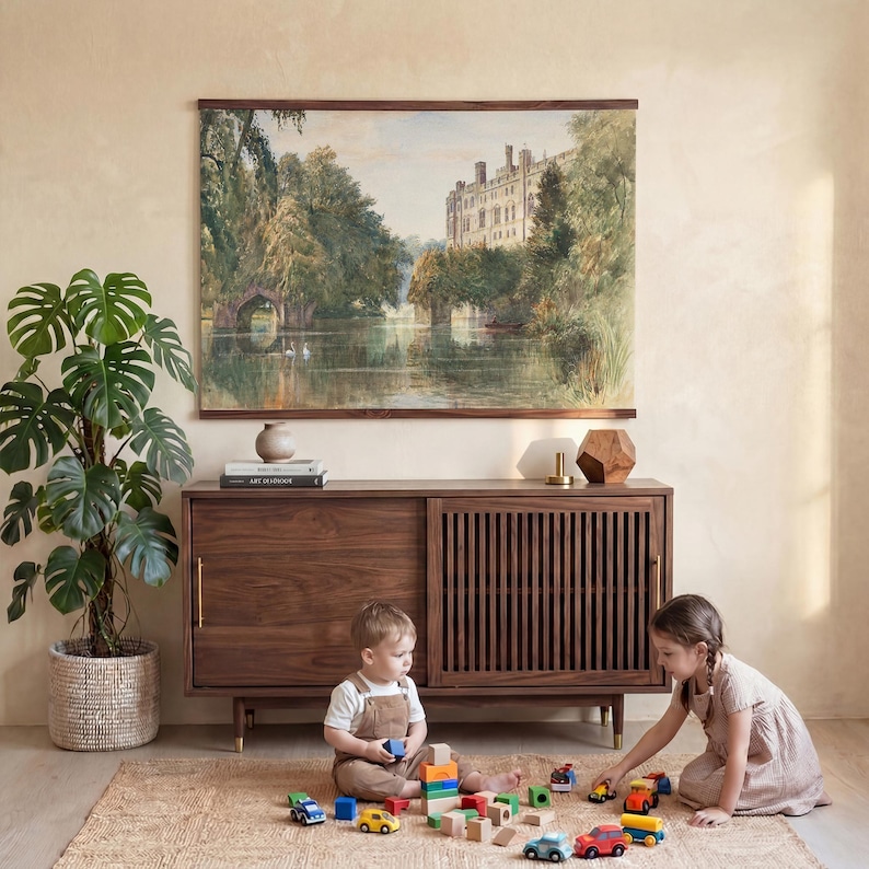 May include: A watercolour print of a castle and river scene hangs above a dark wood cabinet. A large potted plant sits to the left. Two children play with toy cars and blocks on a woven rug in front of the cabinet.