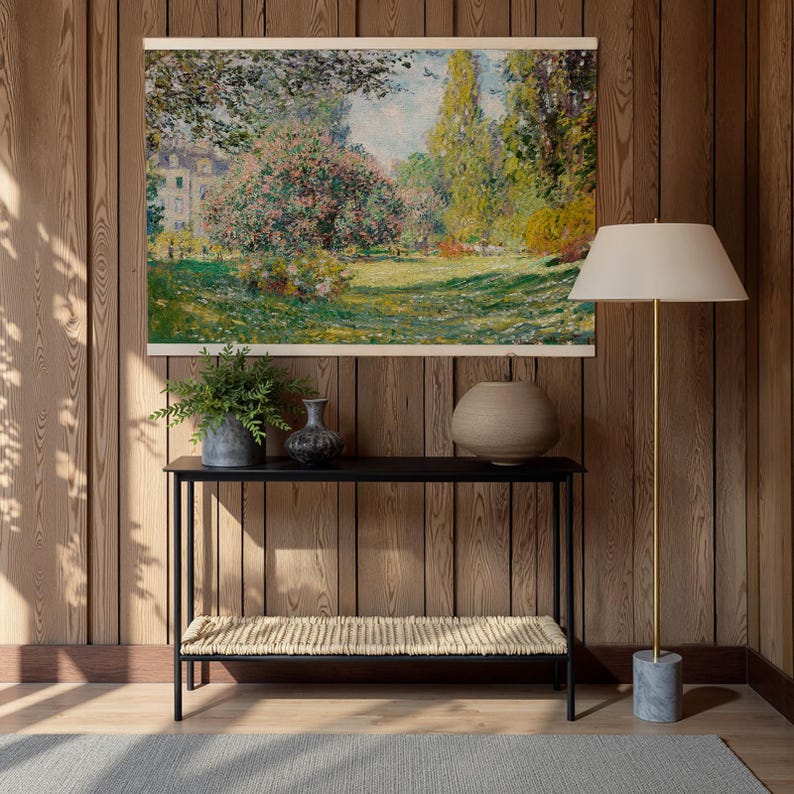 Pode incluir: Uma pintura de paisagem emoldurada retratando uma cena de parque com &aacute;rvores, flores e um edif&iacute;cio ao fundo. Abaixo, uma consola preta com um banco tecido e vasos decorativos. &Agrave; direita, uma lumin&aacute;ria de ch&atilde;o com abajur bege.