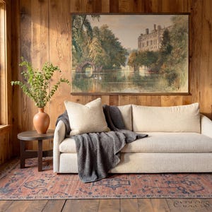 May include: A living room with a cream sofa, grey throw, and beige cushion. A framed landscape painting hangs above the sofa. A dark brown side table holds a terracotta vase with greenery. A patterned rug covers the floor. The room has wooden paneling.