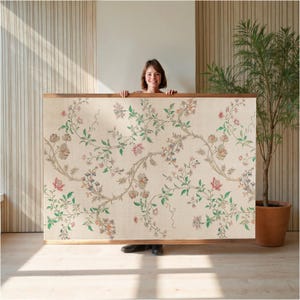 May include: A large rectangular floral tapestry with a wooden frame. The tapestry features a cream-colored background with a repeating pattern of pink, green, and brown flowers and vines. The frame is made of light brown wood.