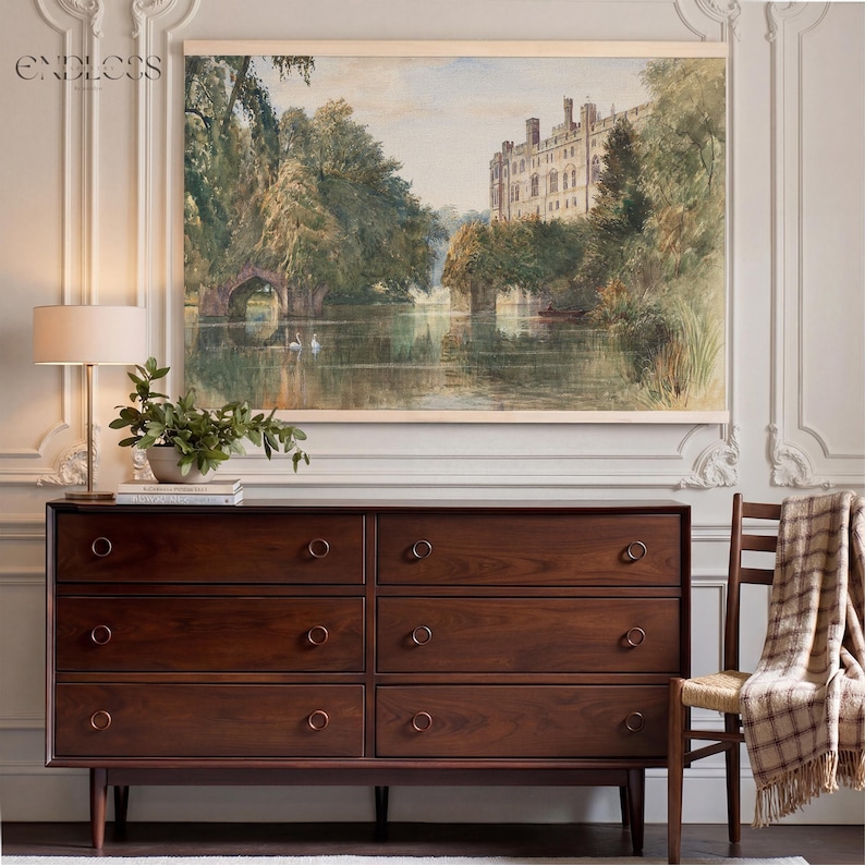 May include: A dark wood chest of drawers with seven drawers and brass ring handles. Above the chest is a framed landscape painting of a castle and a lake. A lamp and a chair with a blanket are also in the image.