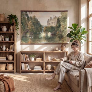 May include: A living room with a large landscape painting of a castle and lake. Wooden bookshelves and a sofa are present. A person is sitting on the sofa reading a book. The room is decorated in neutral tones with natural light.