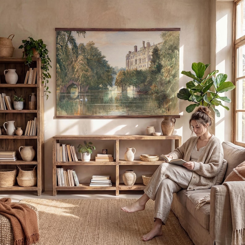 May include: A living room with a large landscape painting of a castle and lake. Wooden bookshelves and a sofa are present. A person is sitting on the sofa reading a book. The room is decorated in neutral tones with natural light.