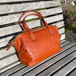 May include: A brown leather tote bag with a zipper closure and two top handles. The bag is sitting on a wooden bench.