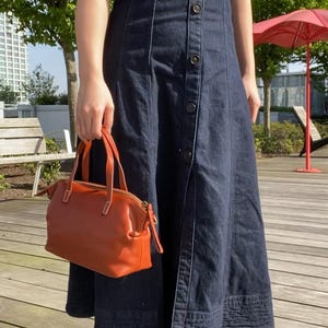 May include: A woman wearing a dark blue denim dress with a button-up front and a brown leather handbag. She is wearing black ankle boots with heels.