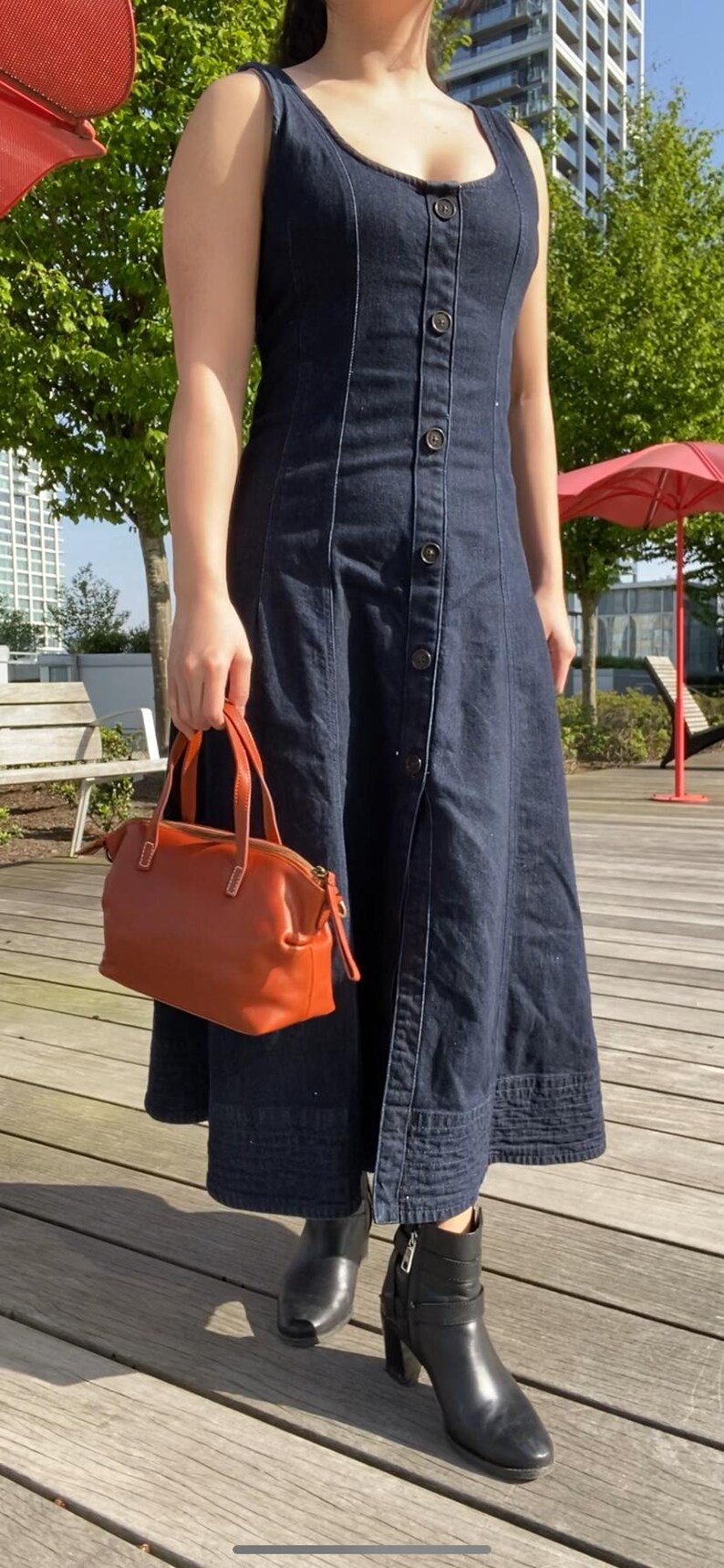May include: A woman wearing a dark blue denim dress with a button-up front and a brown leather handbag. She is wearing black ankle boots with heels.