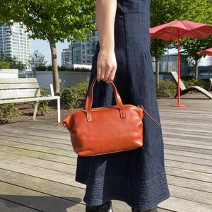 May include: A woman wearing a dark blue denim dress and black heeled boots is holding a brown leather satchel bag with a top handle.