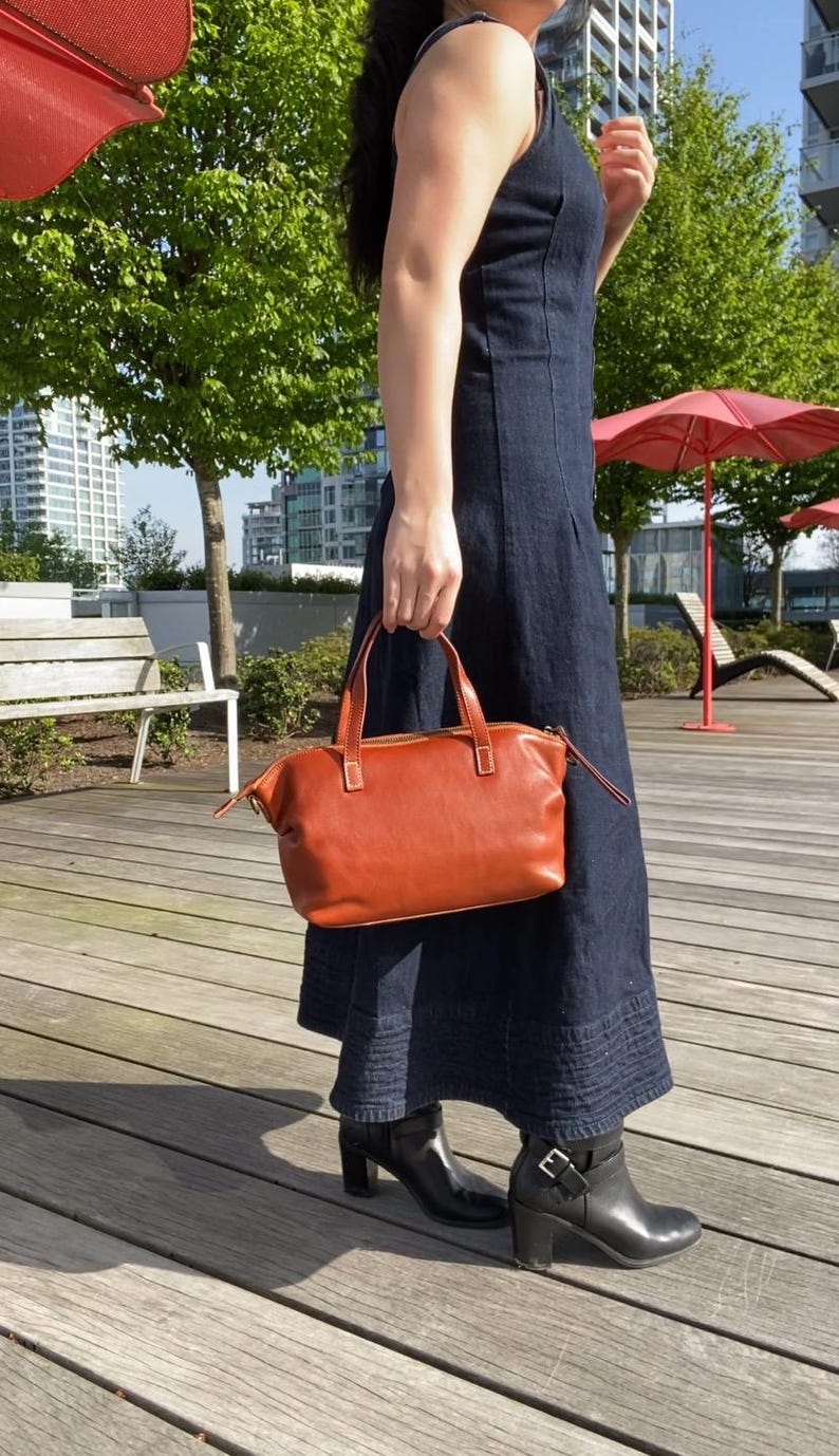 May include: A woman wearing a dark blue denim dress and black heeled boots is holding a brown leather satchel bag with a top handle.