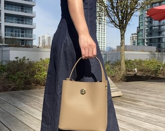 May include: A woman wearing a dark blue denim dress and black heeled boots is holding a tan leather bucket bag with a gold clasp.