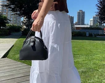 May include: A black leather handbag with a top handle and a buckle closure. The bag has a rounded shape and a structured design. The person is wearing a brown top and a white tiered skirt. The background includes a wooden walkway, green grass, and buildings.
