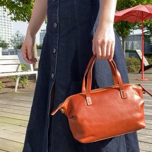 May include: A woman wearing a dark blue denim dress and black heeled boots is holding a brown leather handbag with a zipper closure.