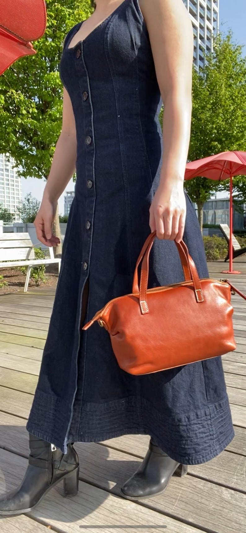 May include: A woman wearing a dark blue denim dress and black heeled boots is holding a brown leather handbag with a zipper closure.