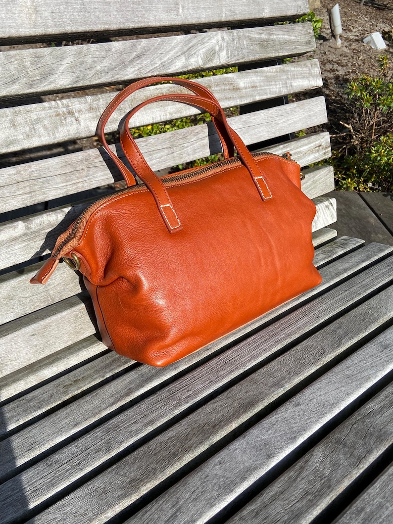 May include: A brown leather tote bag with a zipper closure and two top handles. The bag is sitting on a wooden bench.