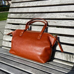 May include: A brown leather tote bag with a zipper closure and two top handles. The bag is sitting on a wooden bench.