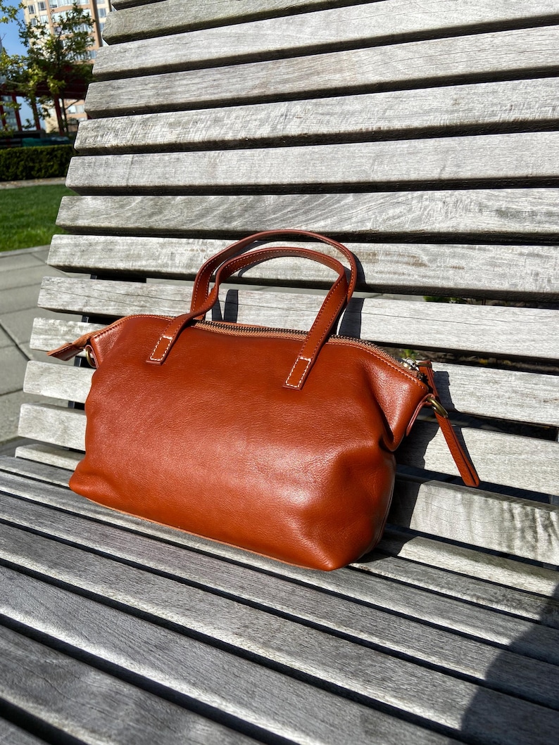 May include: A brown leather tote bag with a zipper closure and two top handles. The bag is sitting on a wooden bench.