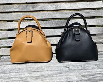 May include: Two leather handbags, one tan and one black, sit side-by-side. Each bag has a rounded shape, a top handle, and a flap closure with a silver buckle. The bags are on a wooden bench.