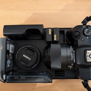 May include: A black Canon camera with a lens attached, a battery pack, and two spare batteries. The camera is sitting on a black surface.