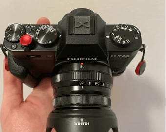 May include: A black Fujifilm X-T20 camera with a black lens attached. The lens has a black lens hood with the Fujifilm logo on it. The camera has a black strap attached to it. The camera has a red button on the top.