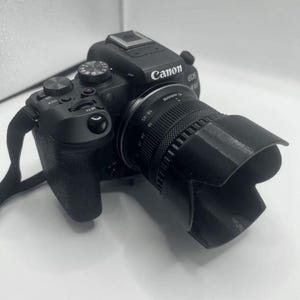 Op de afbeelding: Een zwarte Canon EOS Rebel T7i camera met een lens en een lenskap. De camera heeft verschillende knoppen en draaiknoppen, waaronder een zoomregelaar, een modusknop en een sluiterknop.