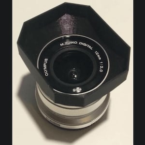 Pode incluir: Uma capa de lente preta está presa a uma lente Olympus M.Zuiko Digital 12mm 1:2.0 prateada.