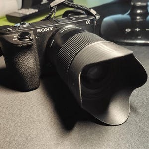 以下が含まれることがあります： 黒いソニーαカメラ、レンズ、レンズフード。カメラには黒いストラップが付いており、暗い場所に置かれています。レンズは黒色で、リブ加工が施されています。