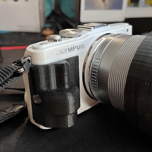 Puede incluir: Una cámara digital Olympus blanca con una lente negra y una empuñadura negra. La cámara tiene una correa negra y el logotipo de Olympus es visible en el lateral. La lente es grande y cilíndrica.