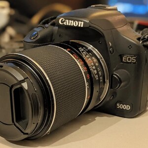 May include: A black Canon EOS 500D digital SLR camera with a black lens attached. The lens has a silver ring with red markings and numbers.